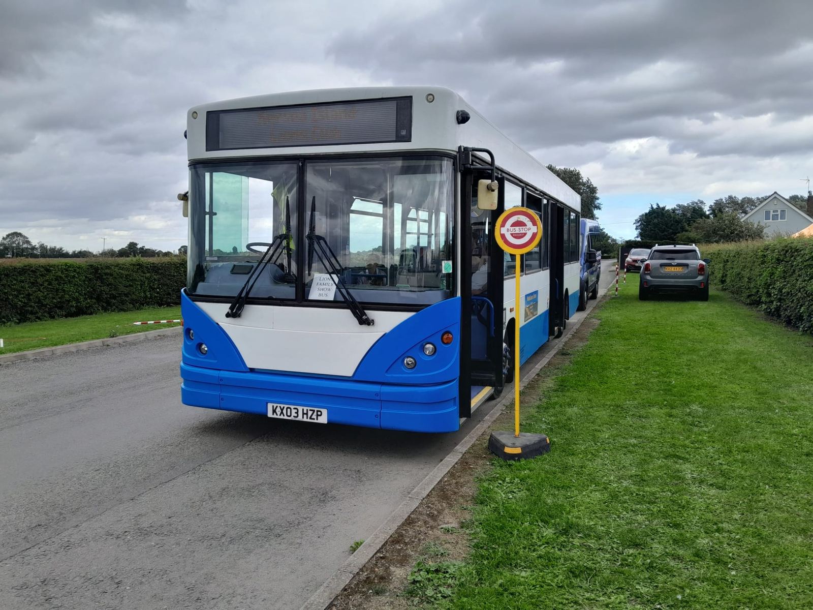 Front of a blue and white bus beside a bus stop pole on a grass verge