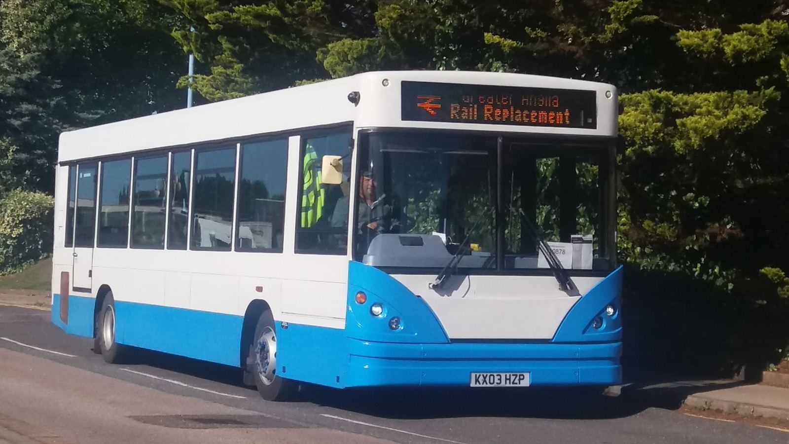 White and blue single-decker bus showing a Rail Replacement destination display, parked beside trees