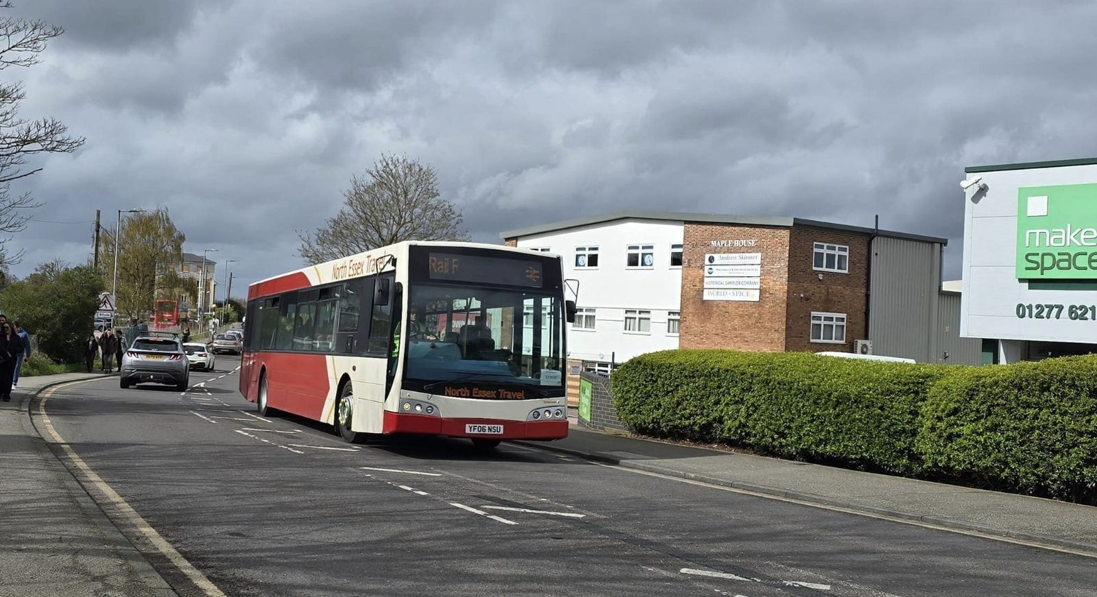 Red and white bus driving along a town road past hedges and offices