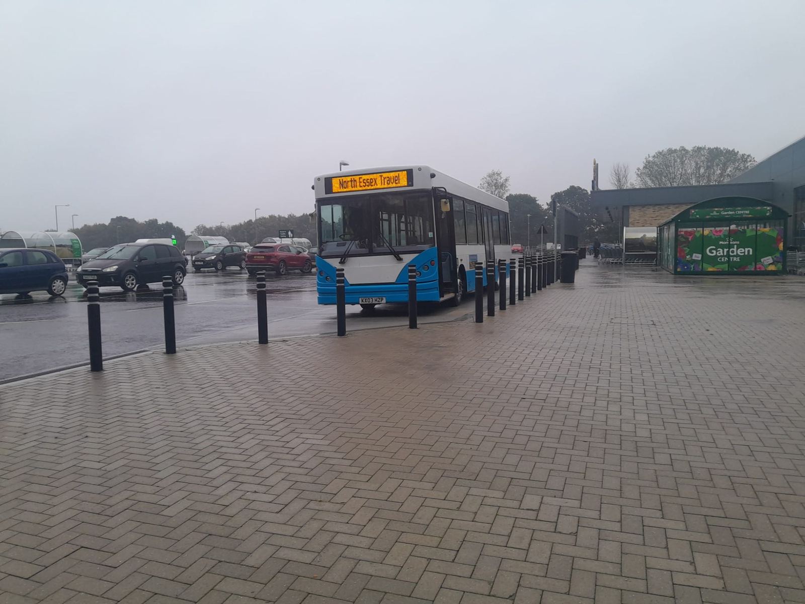 Blue and white bus parked on a wet paved car park beside a garden centre on a grey day