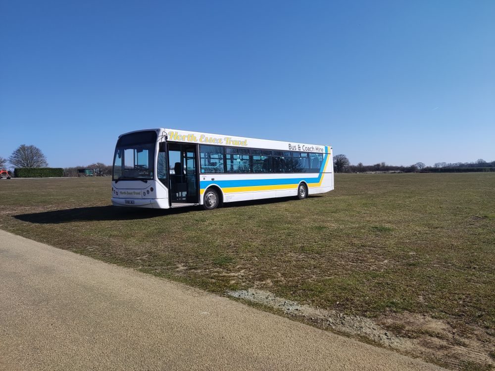 North Essex Travel single-decker bus in white with blue and yellow stripes, parked on grass under a blue sky